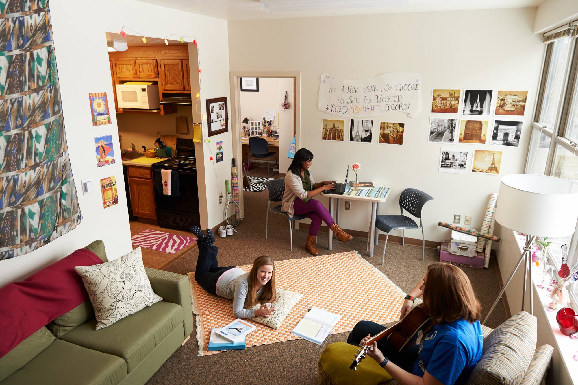 Three people relax in a South E apartment unit. One plays guitar on a sofa, another uses a laptop at a desk, and a third lies on the floor, smiling.